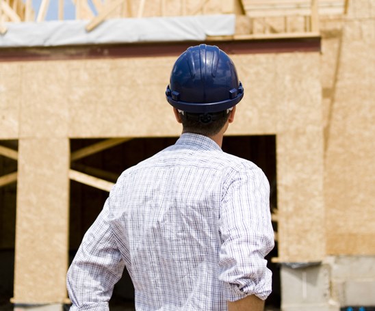 Contractor with back to camera, wearing blue hard hat and button-down collared shirt, looking at building under construction.