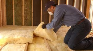 Technician kneeling on unfinished floor, removing yellow fiberglass batt insulation.