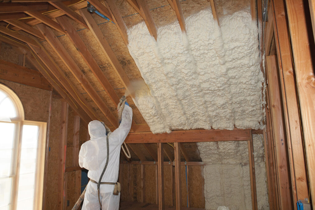 Insulation technician installing spray foam insulation in an attic ceiling while wearing a white hazmat suit.
